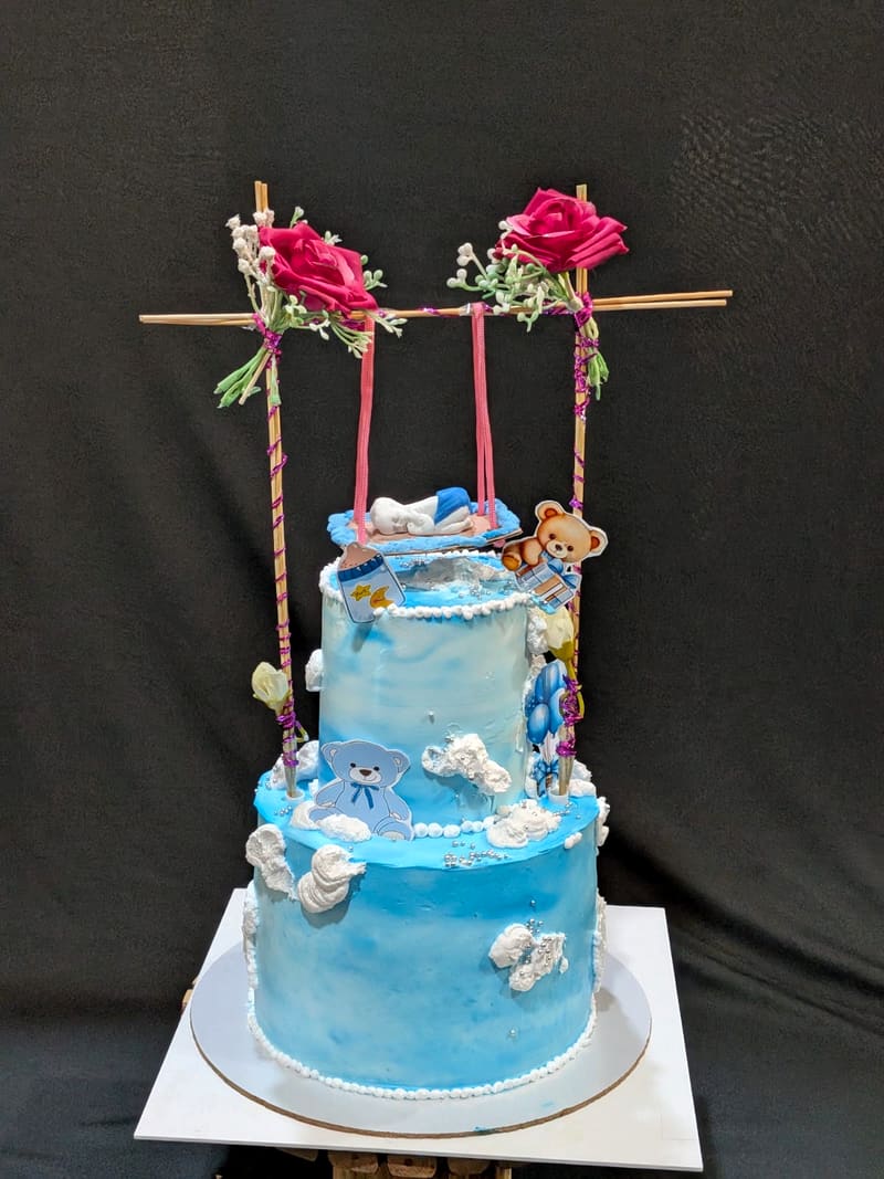 Two-tier blue watercolour-effect baby-shower cake with a bamboo swing structure holding a fondant sleeping baby, deep pink fabric roses, teddy bear illustrations and white piped clouds