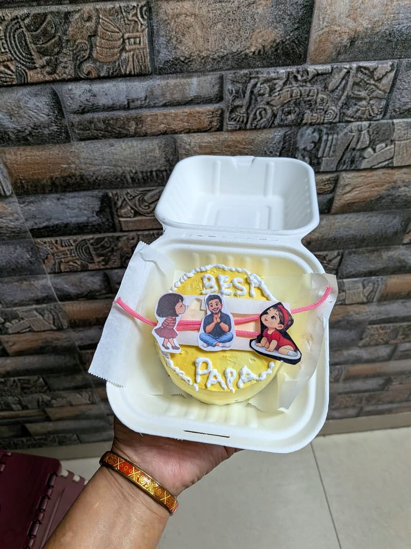 Small yellow bento-style cake in a takeaway box, piped Best Papa lettering, family cartoon cutouts and a pink rakhi thread laid across the top for Raksha Bandhan