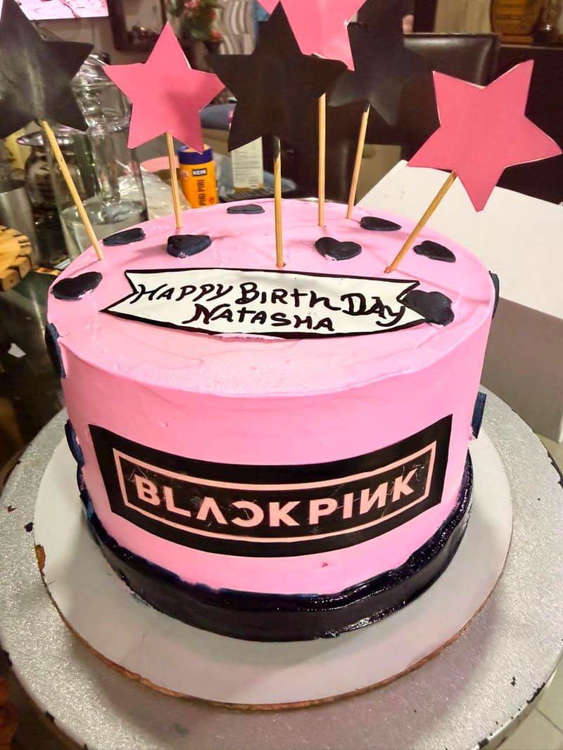 Round pink buttercream cake with a black BLACKPINK logo band, black and pink paper stars on skewers, black fondant hearts, and a hand-drawn banner reading Happy Birthday Natasha