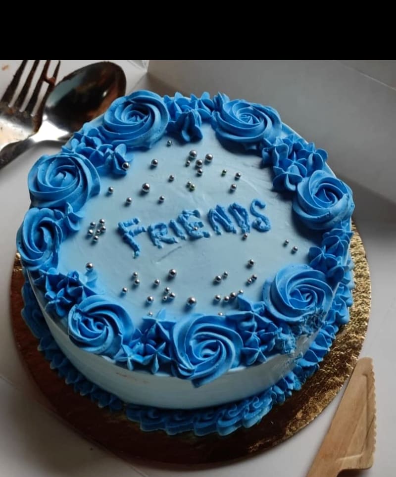 Round cake with a ring of piped blue buttercream roses and star borders, silver dragee sprinkles and a piped hashtag Friends lettering on a light blue base