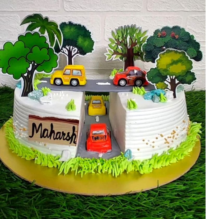 White buttercream cake split by a grey piped road with a bridge overpass, decorated with die-cast toy cars, cut-out paper trees, green buttercream grass, edible rocks and gold sprinkles, with a wood-effect name plaque reading Maharsh