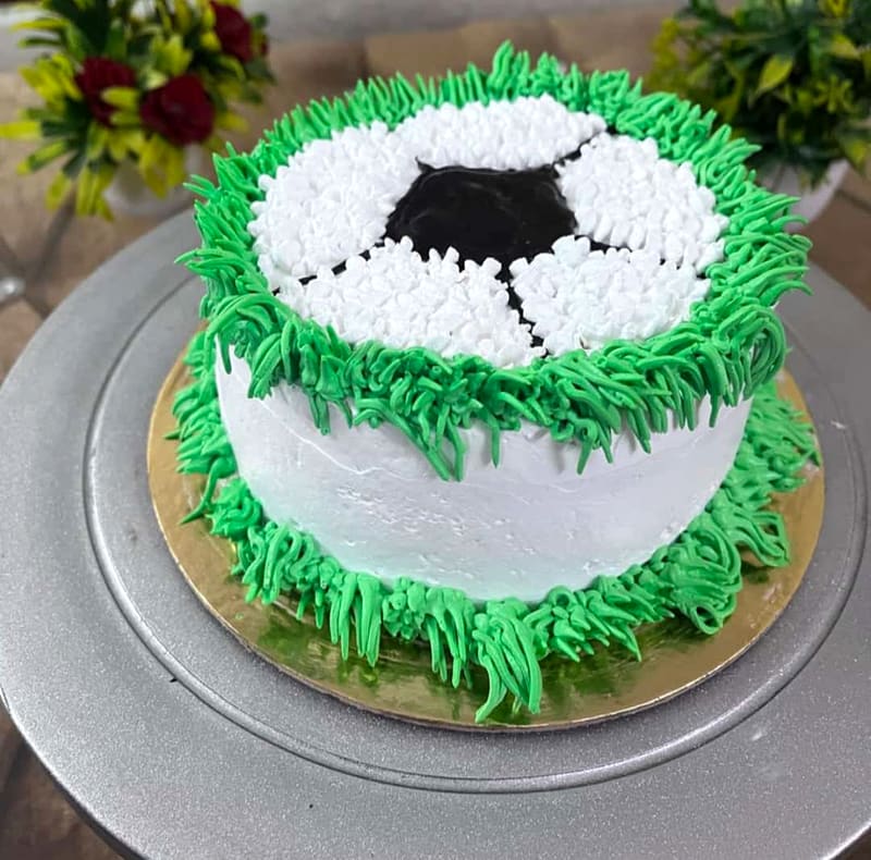 Round cake styled as a football, piped white and black buttercream panels on top ringed with green grass-textured buttercream piping running down the sides, set on a gold cake board