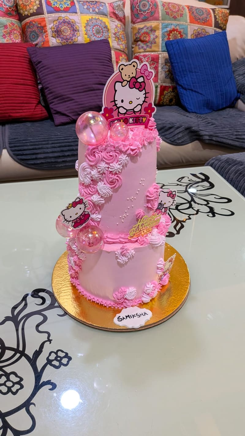 Two-tier pink birthday cake with piped rosettes, white shell borders and iridescent sugar bubbles, decorated with Hello Kitty cutout toppers and a name plaque reading Samiksha