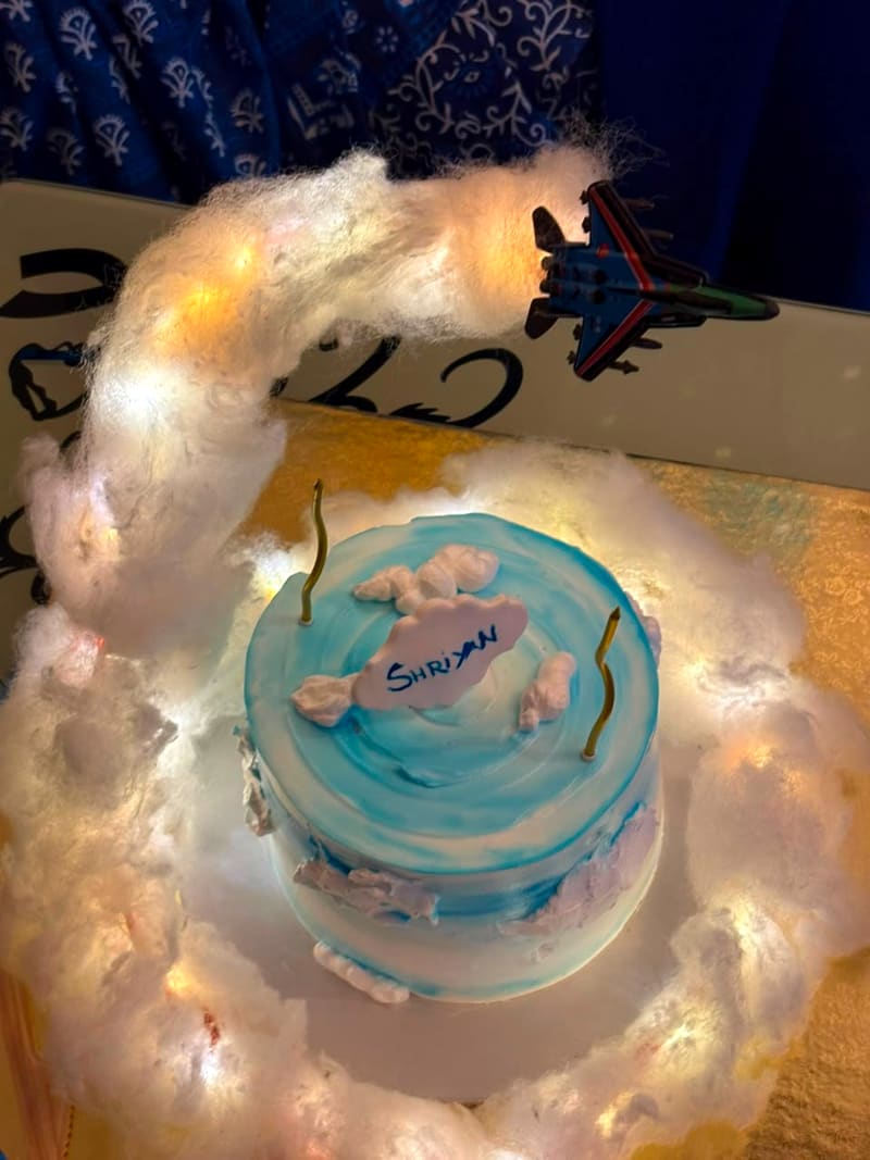 Round blue-and-white buttercream cake styled as sky and clouds, with a die-cast fighter jet posed above a glowing LED-lit cotton-wool contrail and a hand-lettered name plaque reading Shriyan, lit birthday candles on top