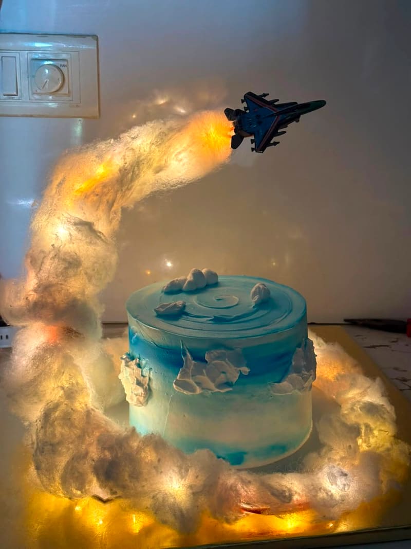 Side view of the same blue sky-and-clouds buttercream cake with a toy fighter jet flying above an LED-lit cotton-wool contrail, cake plain on top without candles or name plaque
