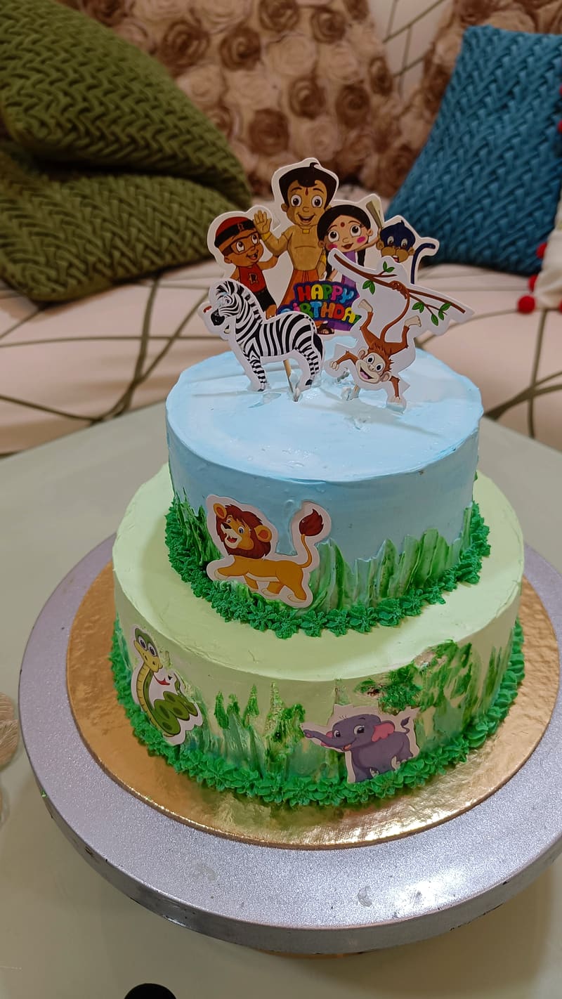 Two-tier jungle-theme cake, sky-blue top tier and pale-green bottom tier piped with grass borders and hand-painted foliage, decorated with printed cutouts of a lion, snake, elephant, zebra and the Chhota Bheem cartoon cast under a Happy Birthday topper