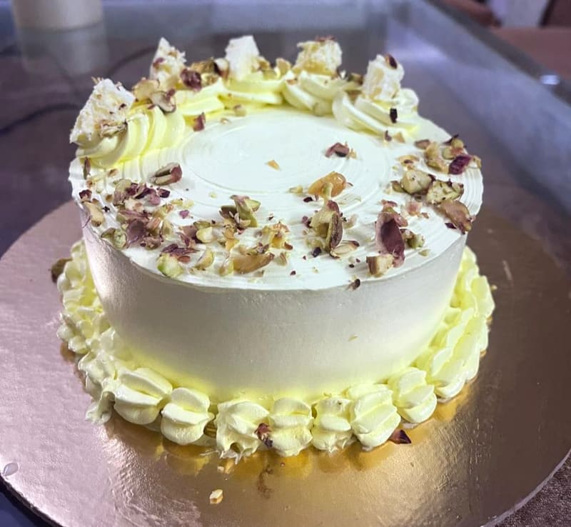 Round cake in pale cream buttercream, piped shell border around the base and rosettes around the top edge, finished with a scatter of chopped pistachios, dried rose petals and shards of wafer on top