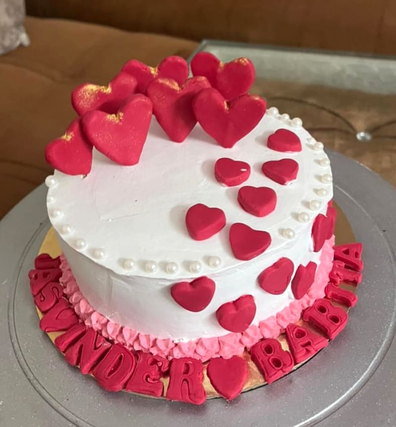 Round white buttercream cake edged with pearl piping and a pink-to-white ombre shell border, scattered with red fondant hearts of varying sizes and finished with the names Sunder and Babita piped in red fondant lettering around the base