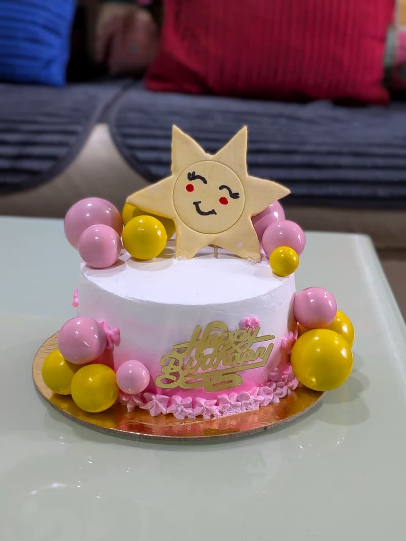 White buttercream birthday cake ringed with pink and yellow sugar spheres, a smiling fondant sun topper, and a gold-script Happy Birthday topper on a pink piped base