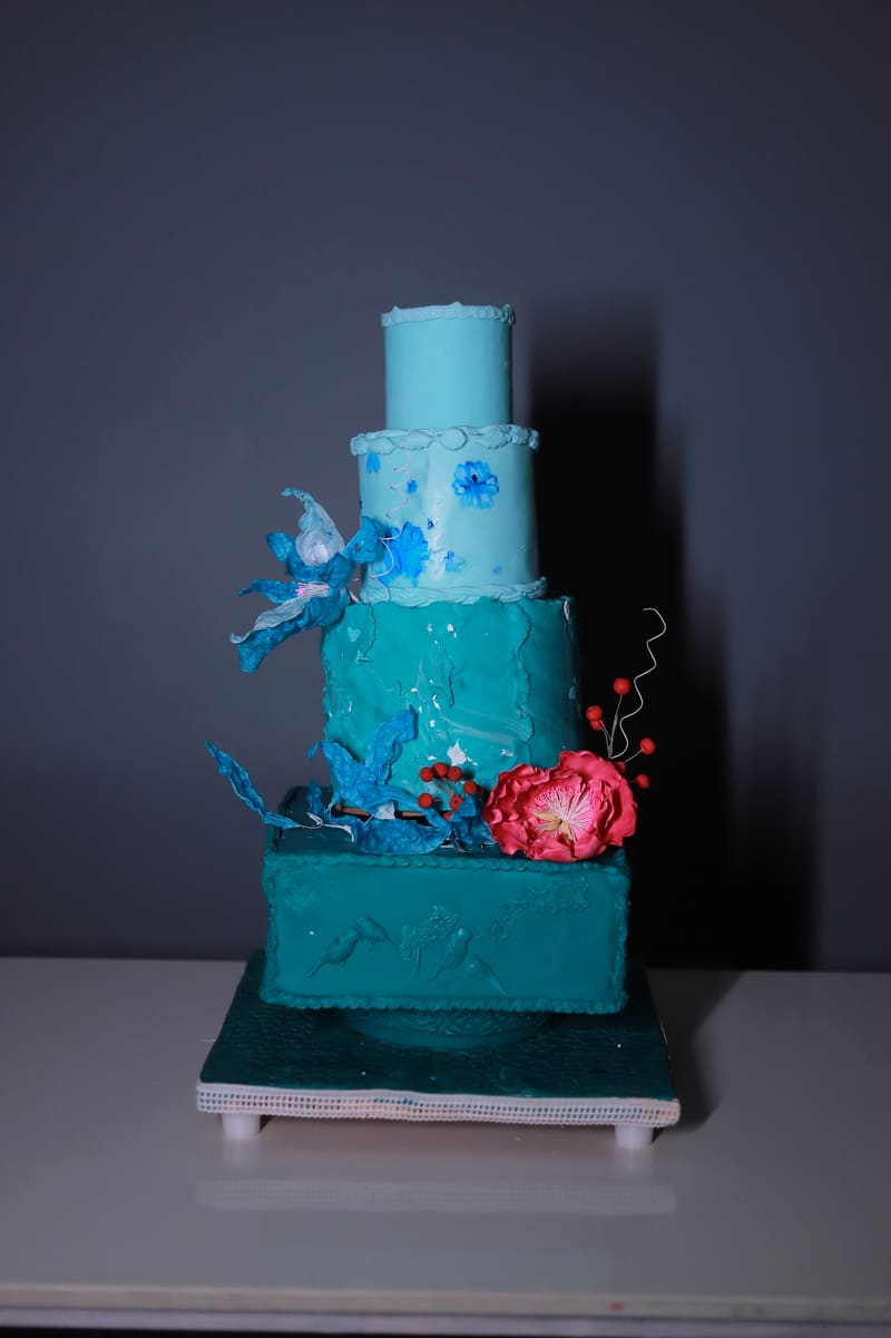 Studio photo on a grey seamless background of a four-tier teal celebration cake: a plain pale-teal cylinder tier, a pale-teal tier hand-painted with small blue flowers, a textured deep-teal tier with a large blue wafer-paper flower and trailing wafer leaves, and a square bas-relief base tier piped with birds and blossoms, finished with a bright pink sugar peony and red berry sprigs