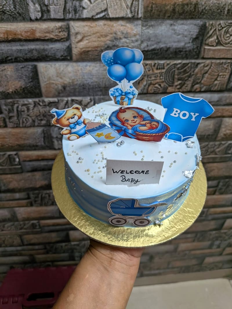 White and blue baby-shower cake topped with a sleeping-baby illustration, teddy bear, blue balloon bouquet and a BOY onesie cutout, edible silver leaf accents and a card reading Welcome Baby