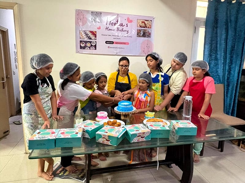 A large group of children in hairnets and aprons gather around Neeta at a glass table to cut a blue piped celebration cake together, teal Neeta's Home Bakery pastry boxes lined up in front of them and the bakery banner on the wall behind