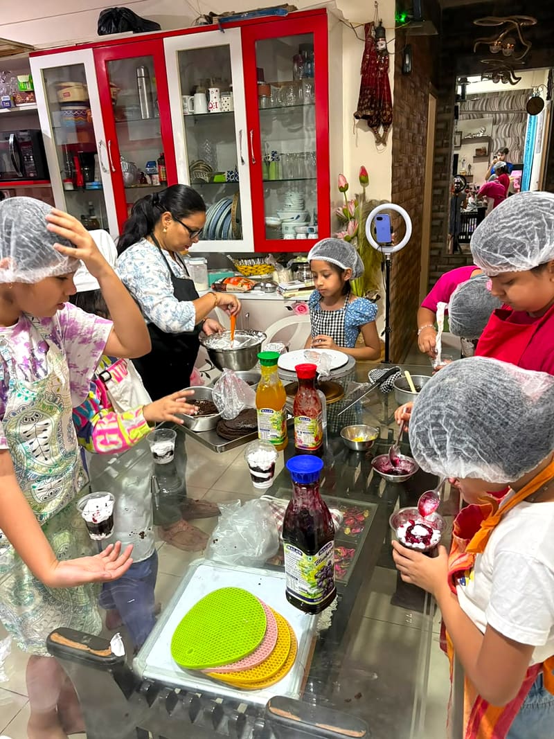 Neeta and a large group of children in hairnets and aprons each hold up a teal Neeta's Home Bakery pastry box packed with their baking, standing together under the bakery banner with its order-now phone number
