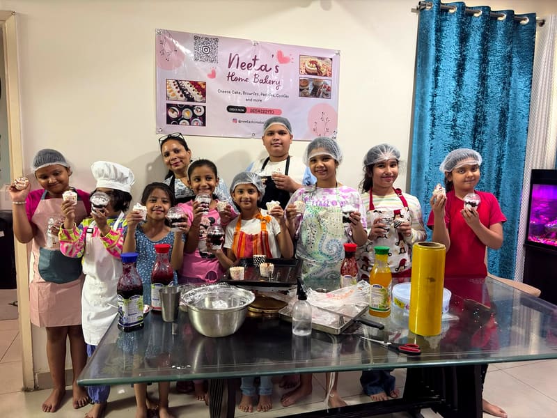 A large group of children in hairnets hold up their own cream-topped cupcakes in front of the Neeta's Home Bakery banner, baking ingredients and sauce bottles spread across the table in front of them