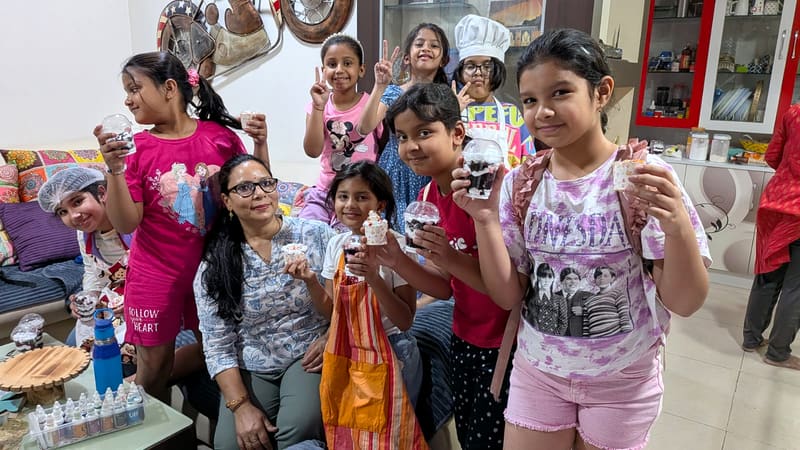 Neeta seated with five children after a home baking class, the children wearing hairnets, aprons and a chef hat and holding their own jar cakes