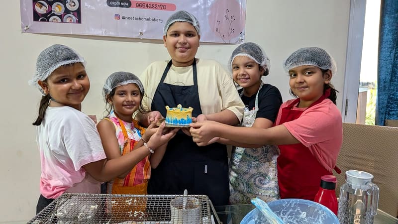Five children in hairnets hold up a small blue-and-white cake they decorated together, standing in front of a Neeta's Home Bakery order-now banner