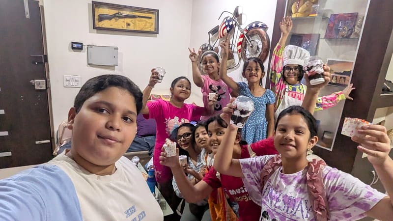 A boy takes a wide selfie with a large group of children behind him, each holding a jar cake topped with cream after a home baking class session