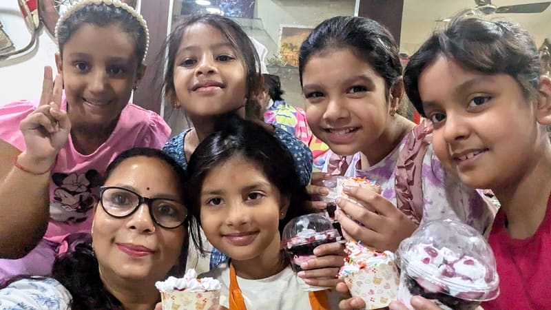 Neeta poses for a selfie with five children after a home baking class, each of them holding a jar cake or cupcake topped with cream and sprinkles