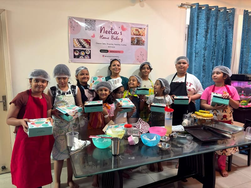 Neeta pipes whipped cream into a piping bag while children in hairnets and aprons gather closely around the table to watch and help, a chandelier and TV unit visible in the living room behind them