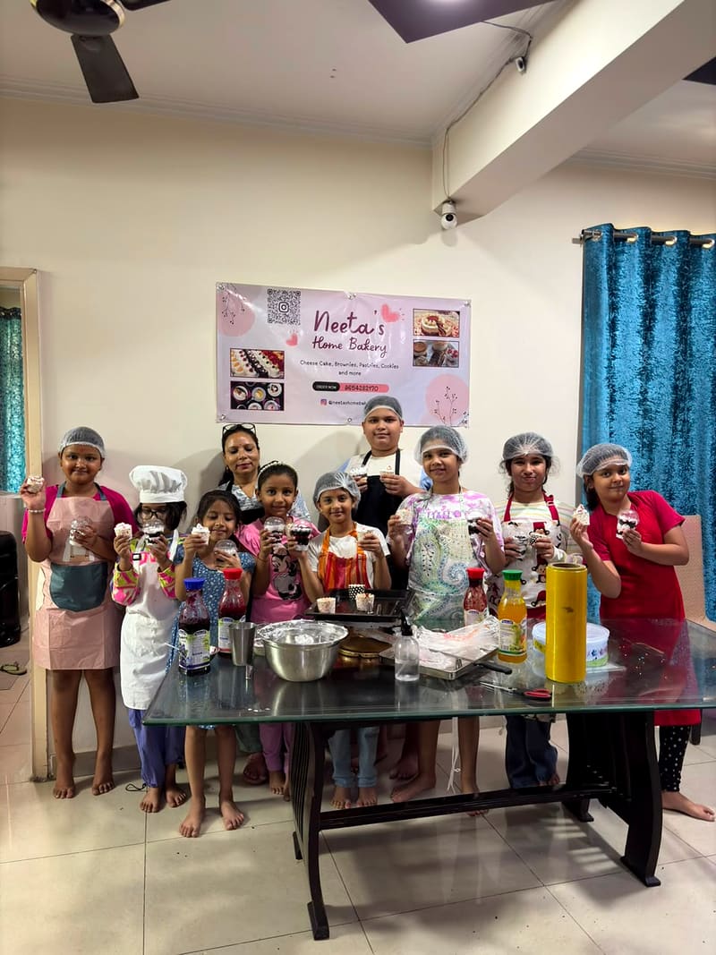 Children in hairnets and aprons crowd around a glass worktable spooning cream and fruit syrup into jar cakes while Neeta helps portion the cream, sauce bottles and mixing bowls spread across the table in front of them