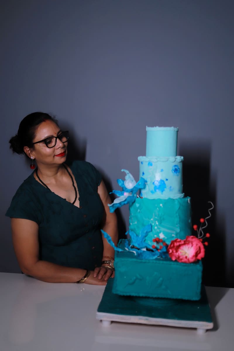 Neeta Srivastava seated at a table beside her four-tier teal celebration cake, looking toward the cake