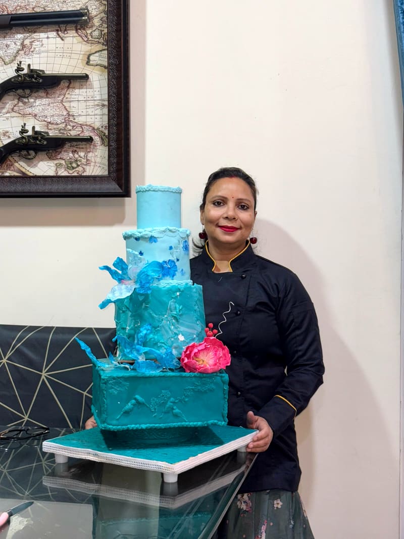 Neeta Srivastava, in a black chef's jacket with gold trim, standing beside her four-tier teal celebration cake and smiling at the camera