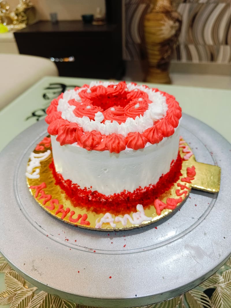 A round red velvet cake finished in white cream-cheese-style frosting, piped in a red-and-white rosette ring on top with a peek of deep red crumb at the centre, and finished with a loose red velvet crumb border around the base